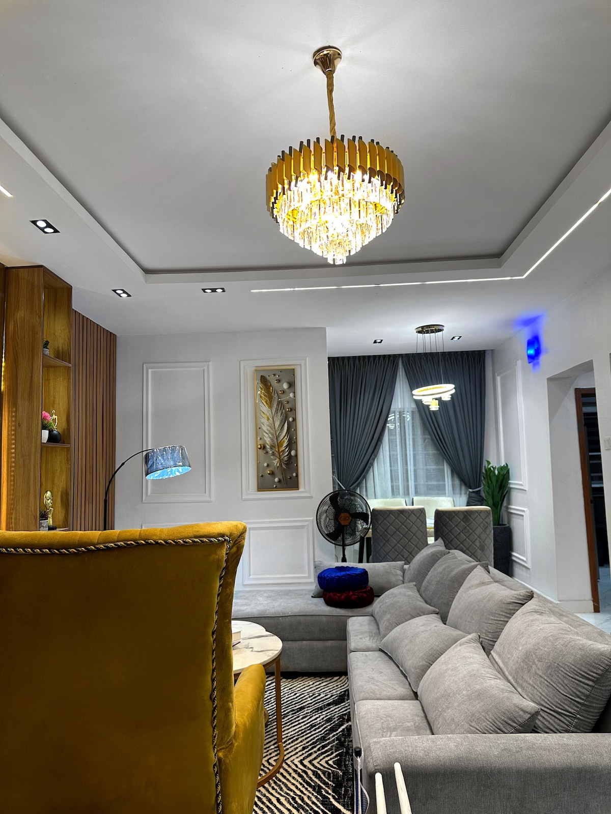 现代客厅，配备电视墙、灰色毛绒组合沙发、金色天鹅绒扶手椅、时尚枝形吊灯、内置木架和时尚咖啡桌。 这里是放松身心、观看节目或招待客人的完美空间，既舒适又时尚。