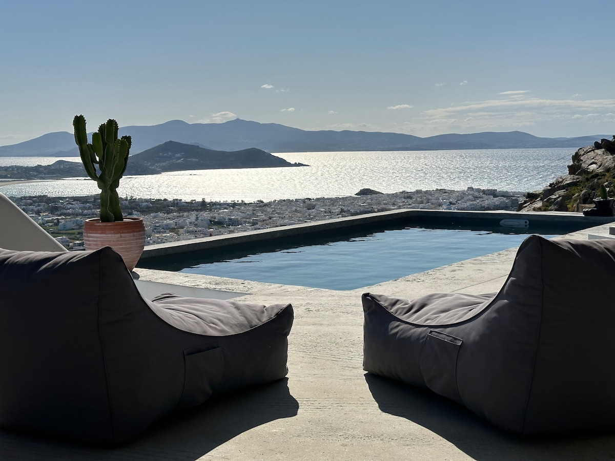 A serene view is captured from a poolside area, featuring two oversized lounge cushions. A potted plant is placed nearby, while the calm waters of Naxos Bay shimmer under sunlight. The panoramic landscape of hills and the town of Naxos create a tranquil background.