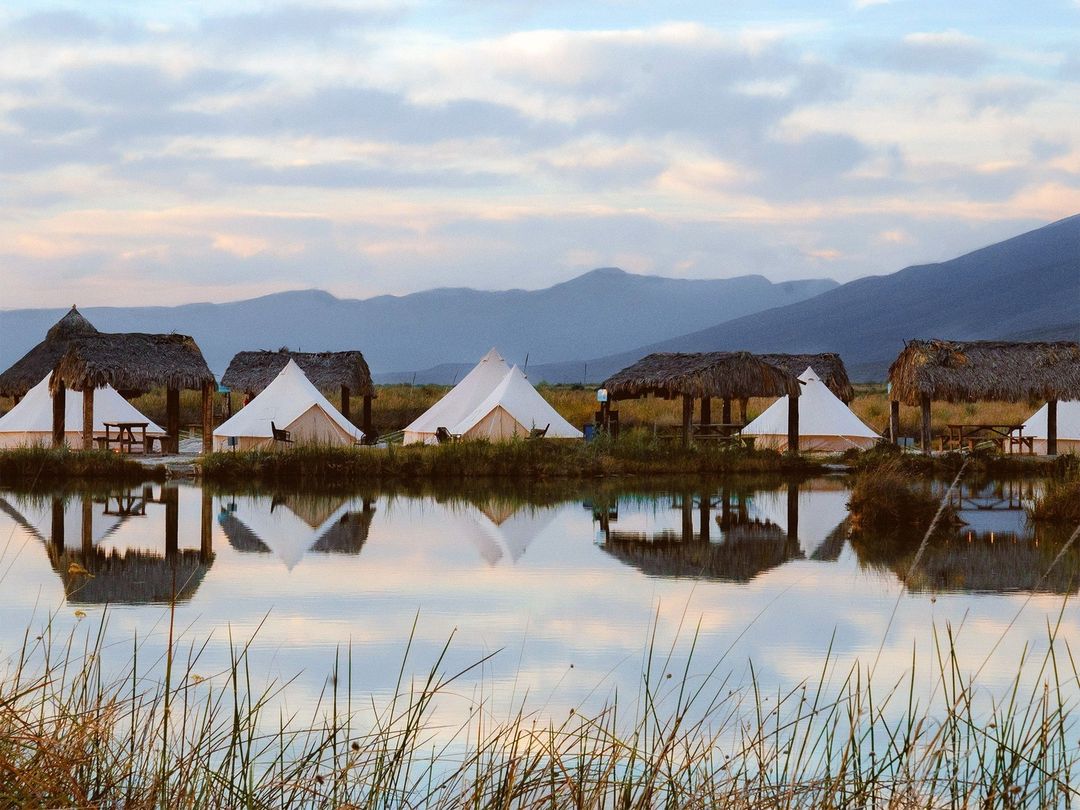 The image depicts several glamping tents arranged near a calm body of water, with reflections mirrored on the surface. Thatched-roof structures are present in the background, alongside a scenic backdrop of rolling hills under a softly lit sky.