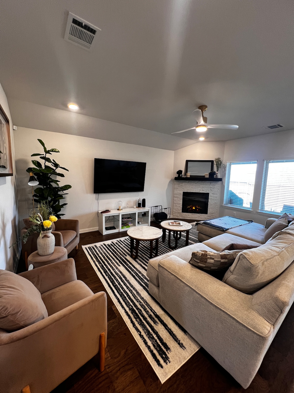 A cozy living area is featured, showcasing a large sectional sofa and two upholstered chairs. A television is mounted on the wall above a modern console. A decorative rug adds texture, while natural light filters through multiple windows, highlighting the fireplace in the corner.
