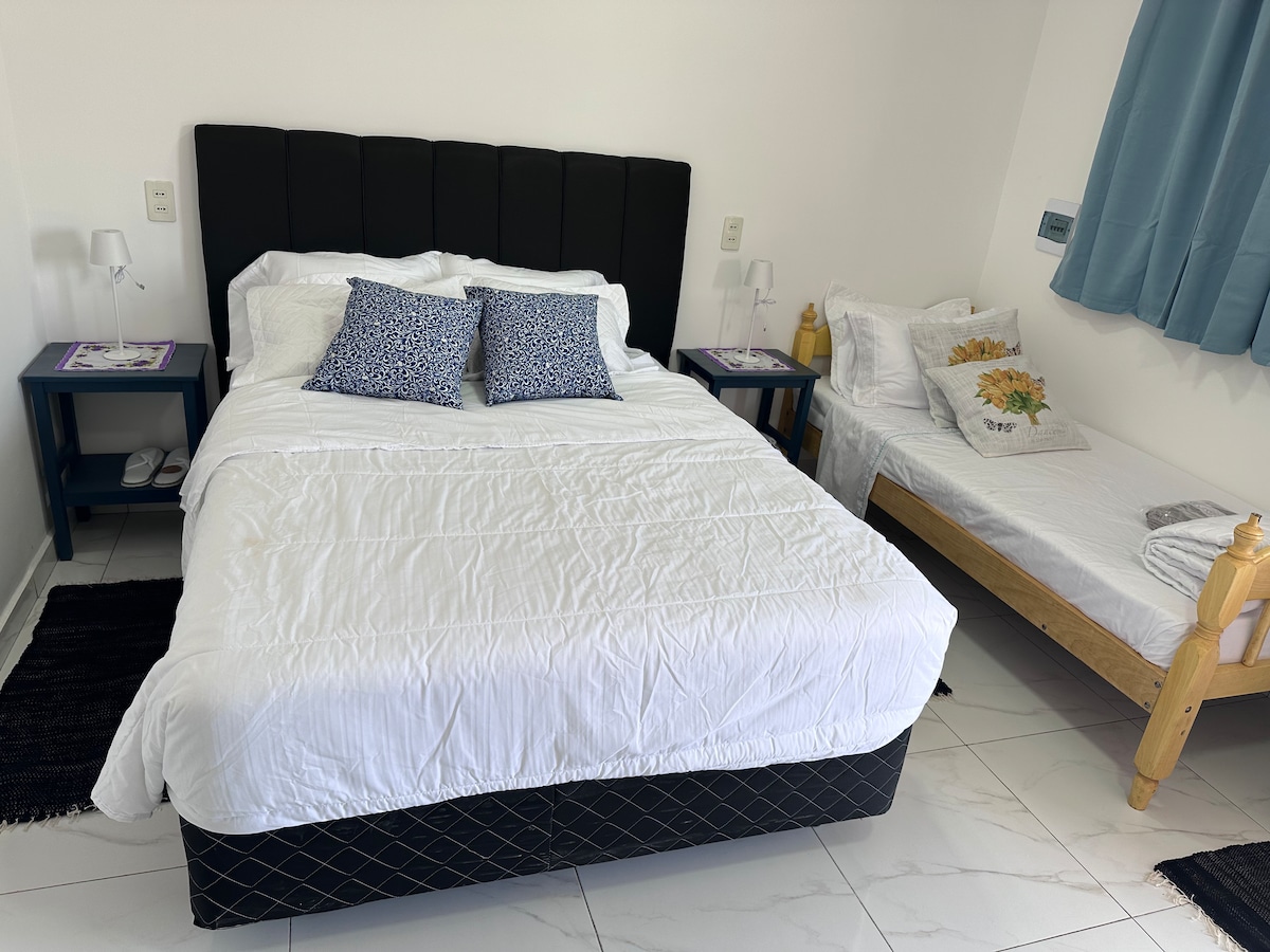 A spacious bedroom is presented with a large bed featuring crisp white linens and two decorative pillows. A smaller single bed is positioned nearby, accompanied by a wooden frame. Soft lighting is provided by modern bedside lamps on blue nightstands. Light blue curtains frame a window.