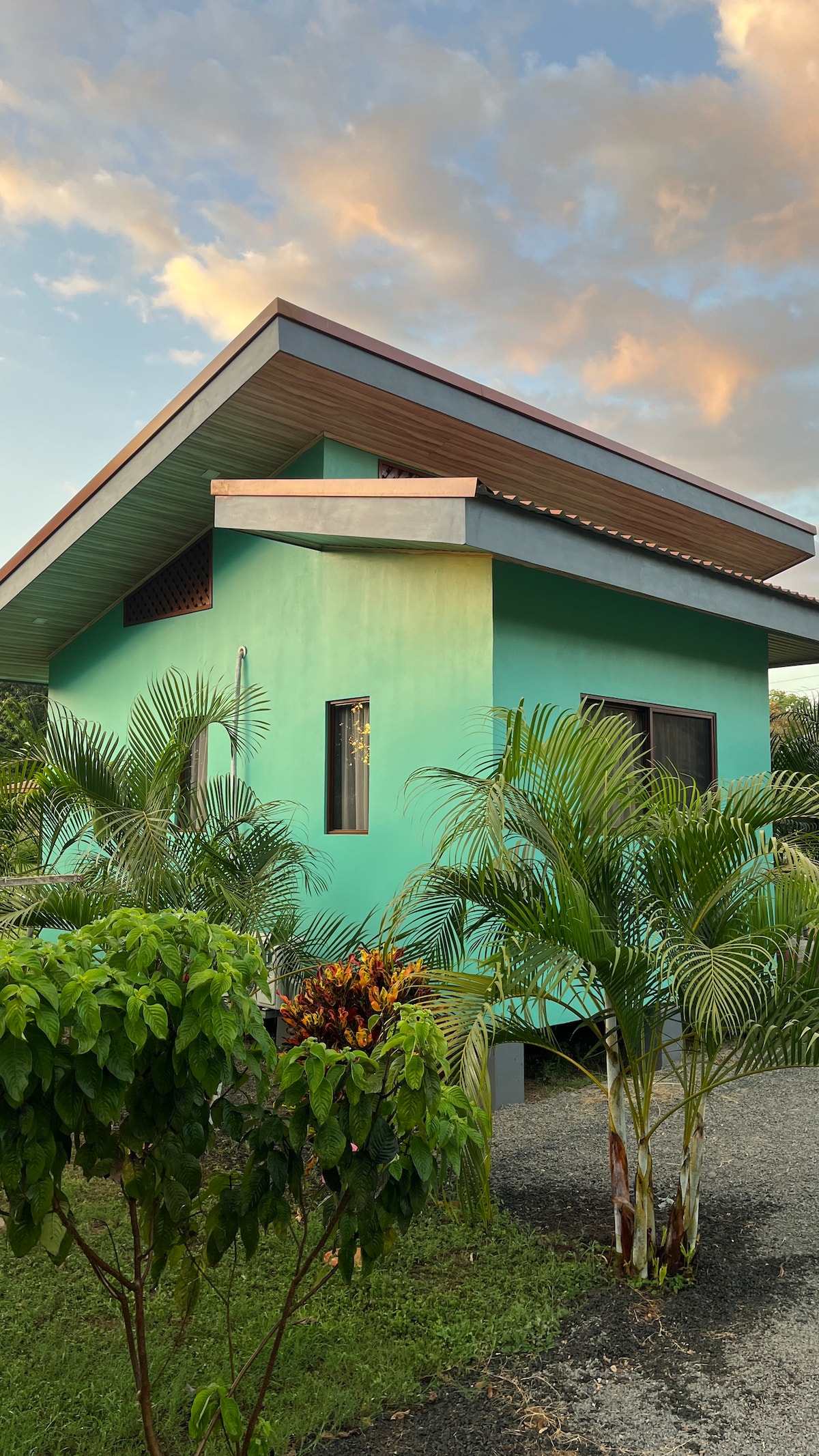 A modern cabin features vibrant turquoise walls under a sloping roof. Large windows are visible, allowing natural light to enter. Lush greenery surrounds the structure, enhancing its tranquil setting. A clear sky with soft clouds is seen in the background.