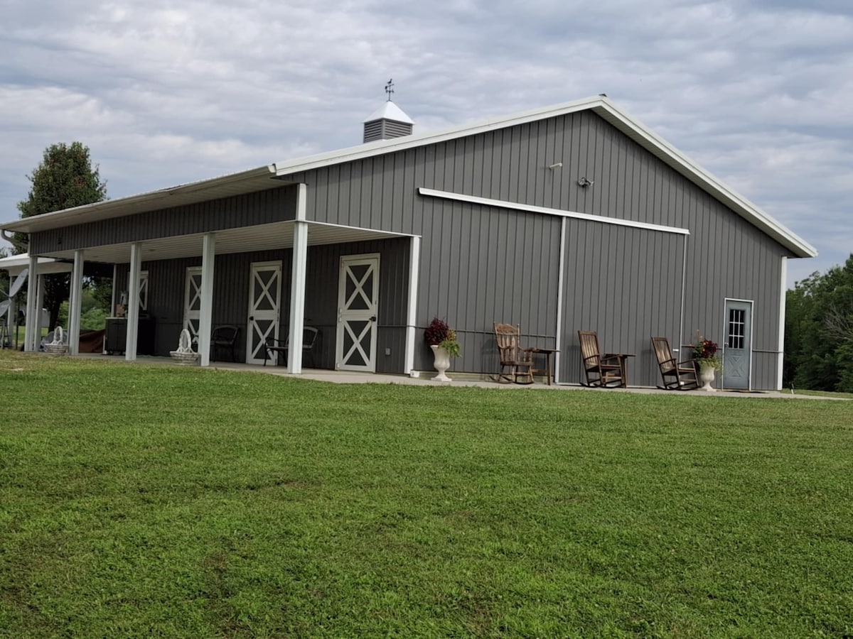 A spacious exterior view of a gray barn-like structure is displayed, featuring stable doors and a wide lawn area. Several rocking chairs are positioned on the covered porch, surrounded by decorative planters. The sky presents soft gray clouds above, enhancing the serene atmosphere.
