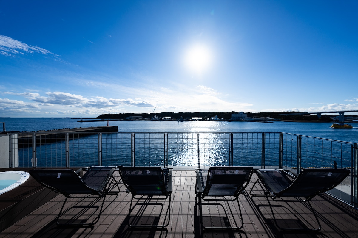A rooftop terrace features four lounge chairs arranged in a row, facing a serene view of the ocean. The sunlight reflects off the water, creating a bright atmosphere. The distant coastline and bridge are visible under a clear blue sky.