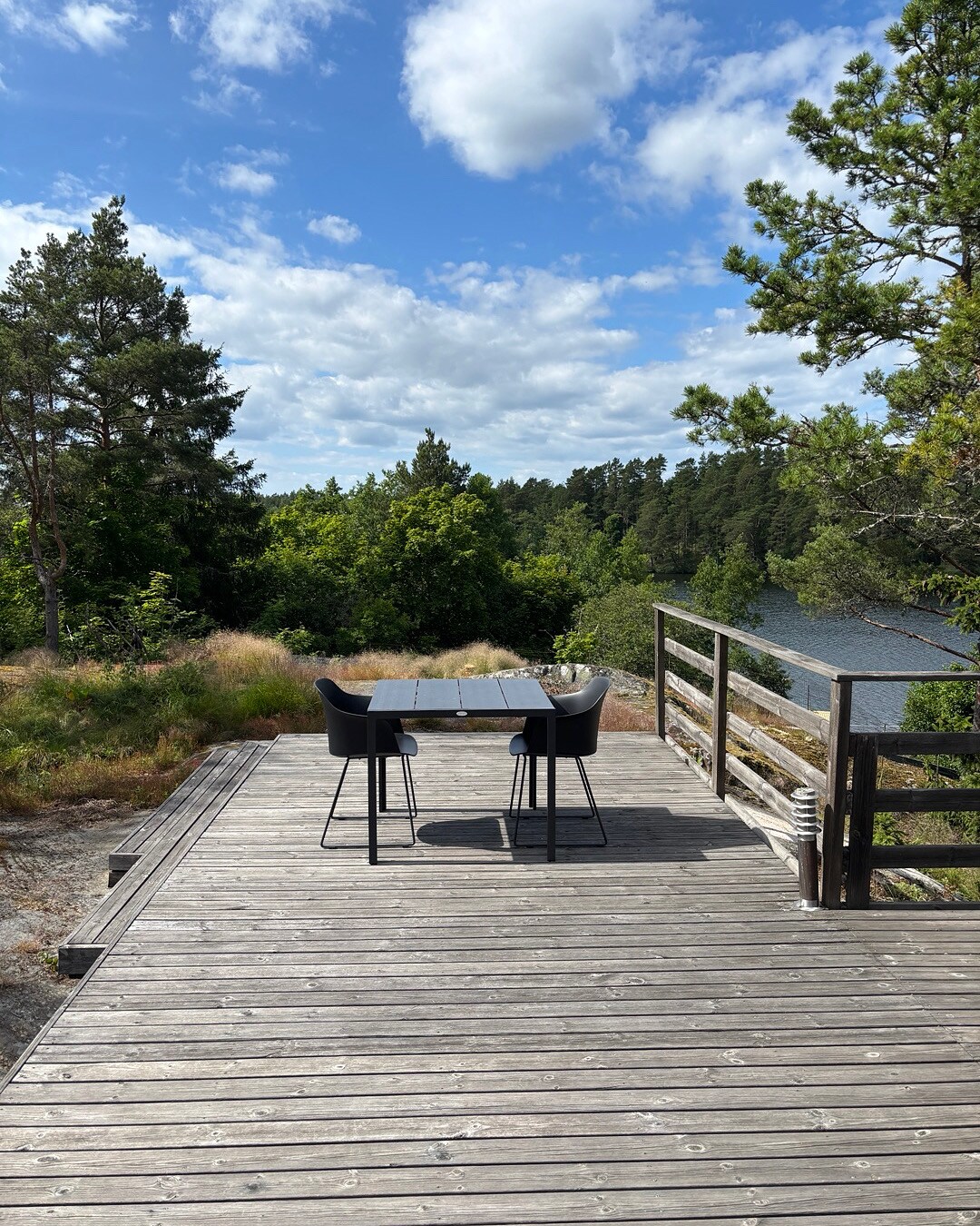 A wooden deck overlooks a serene landscape, featuring a view of lush greenery and a body of water. A simple outdoor dining table with four chairs is positioned on the deck, providing a space for relaxation and enjoyment of the surrounding nature.