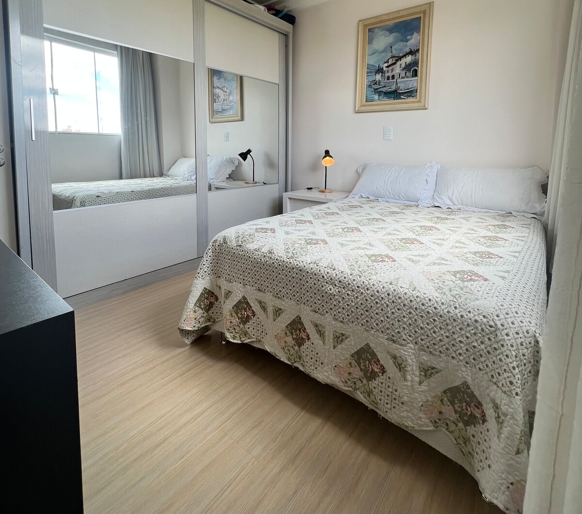 A well-lit bedroom features a spacious bed covered with a patterned blanket. A large mirror reflects the room’s decor, and soft natural light filters through a nearby window, highlighting the neutral color palette. A bedside lamp adds functionality and warmth to the space.