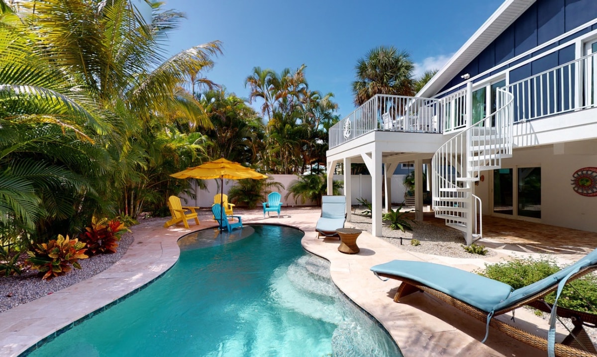 A private heated pool is set within a lush yard, surrounded by tropical plants and palm trees. Colorful lounge chairs and an umbrella provide comfort in the sun. A spiral staircase leads up to a balcony that overlooks the pool area.