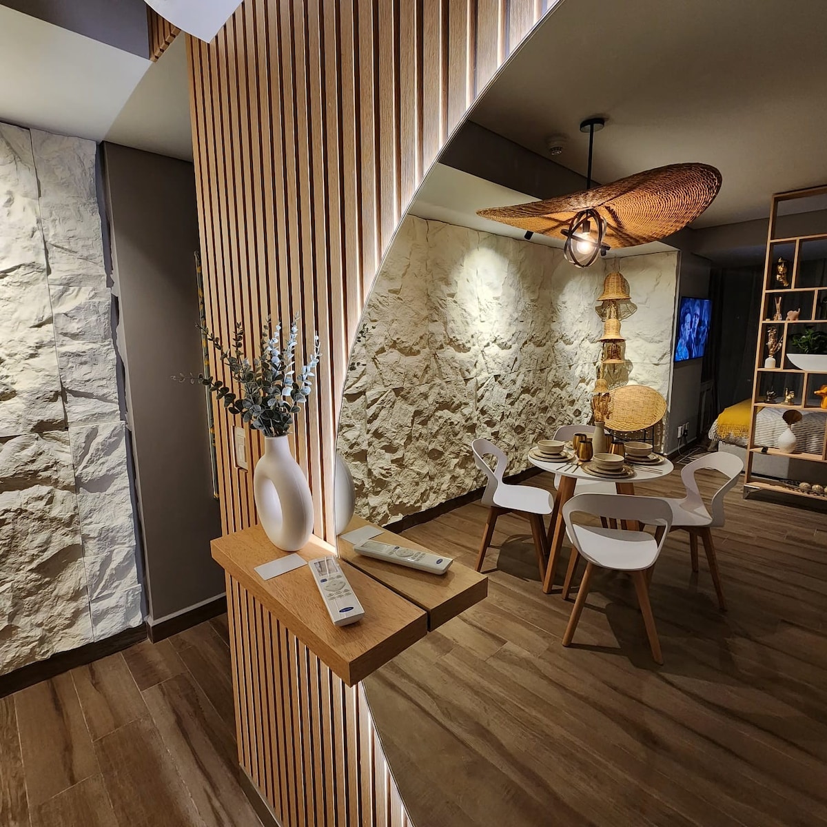 A modern dining area is highlighted by a round wooden table surrounded by four white chairs. Textured stone walls provide a natural backdrop, while a decorative light fixture hangs above. A minimalist shelf displays a vase and other decorative items, enhancing the inviting space.