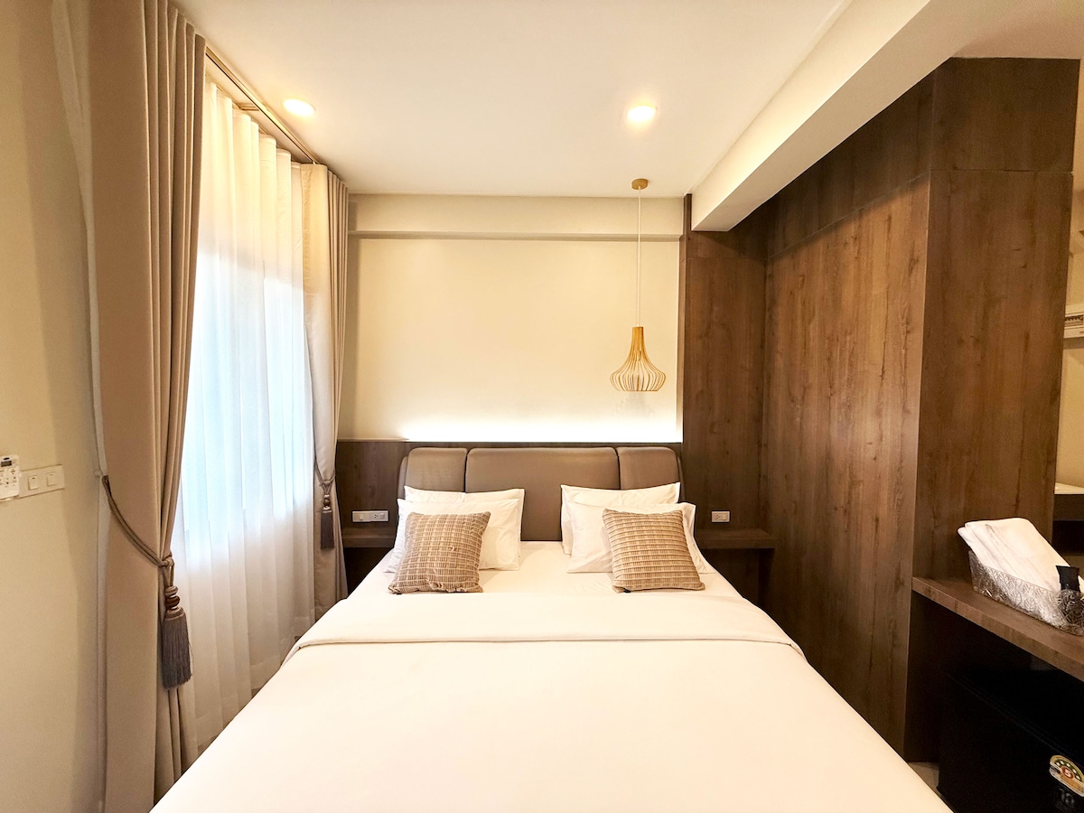 A well-appointed sleeping area features a spacious bed with neutral-colored linens, accented by two decorative pillows. Soft natural light enters through a window dressed in sheer curtains, while a wooden wall complements the modern design. A bedside lamp adds a touch of warmth.