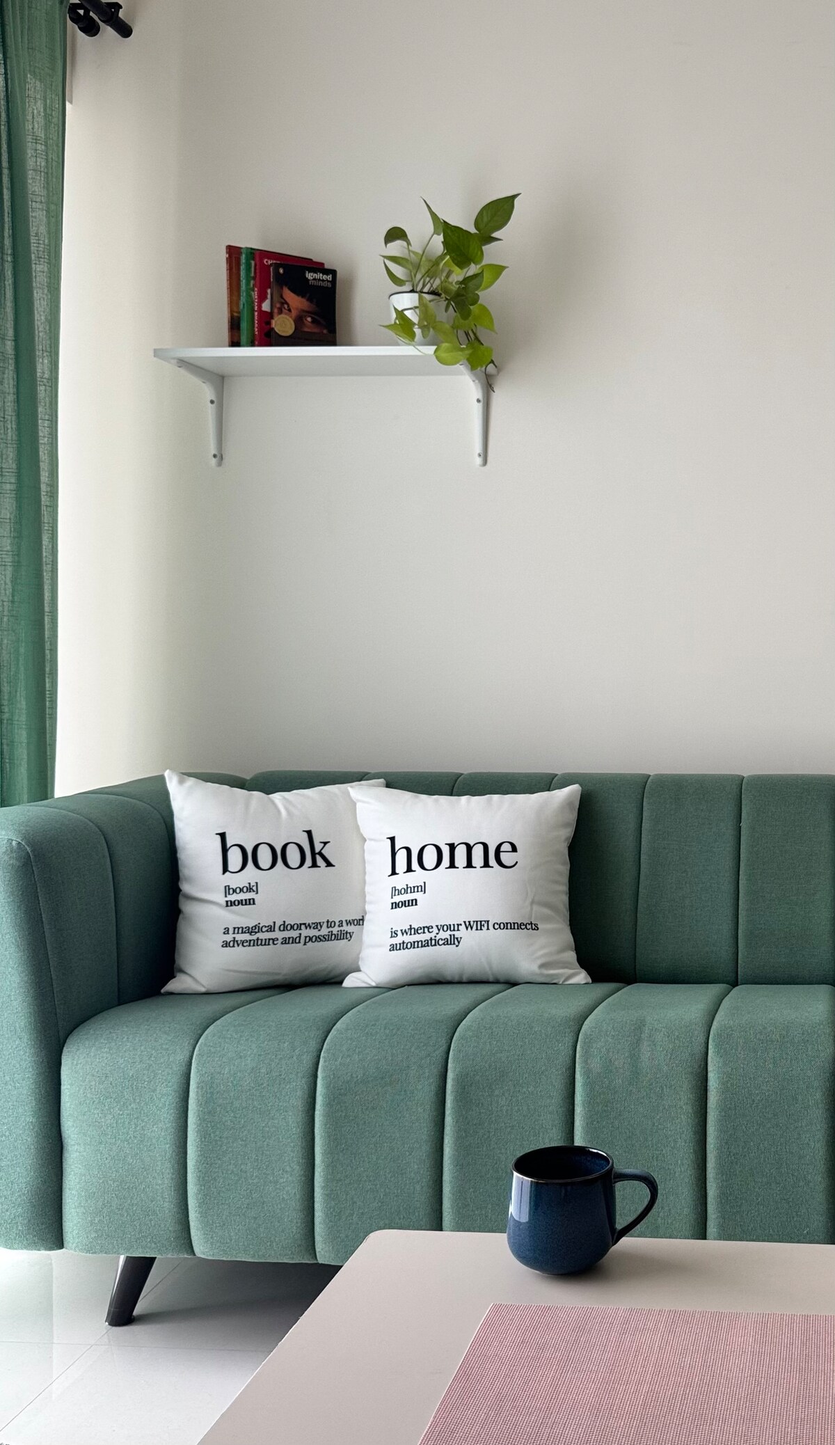 A cozy seating area features a soft green couch adorned with two decorative pillows that emphasize the words 'book' and 'home.' Above, a shelf holds a couple of books and a small potted plant, adding a touch of greenery to the space.