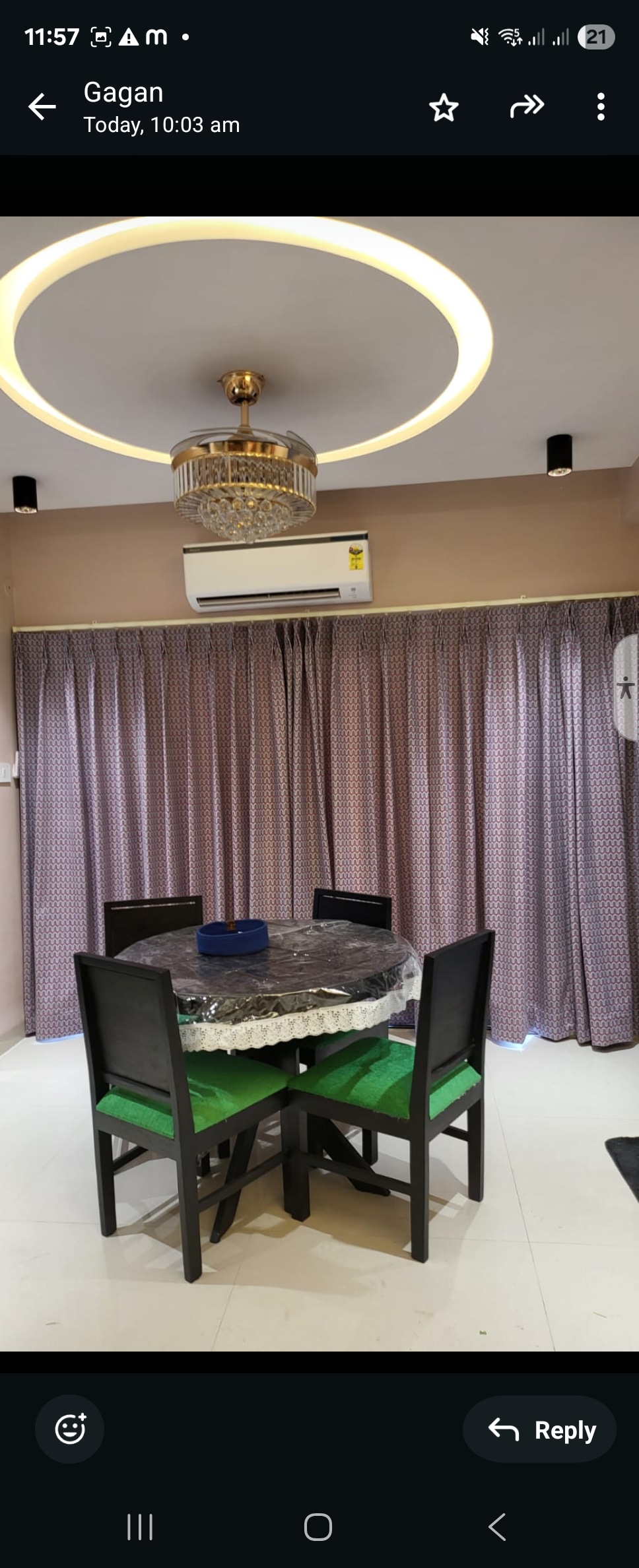 A dining area features a round table surrounded by four chairs, two of which are accented in green. A ceiling light with a decorative design illuminates the space, while soft curtains hang in the background. An air conditioning unit is mounted above the table.
