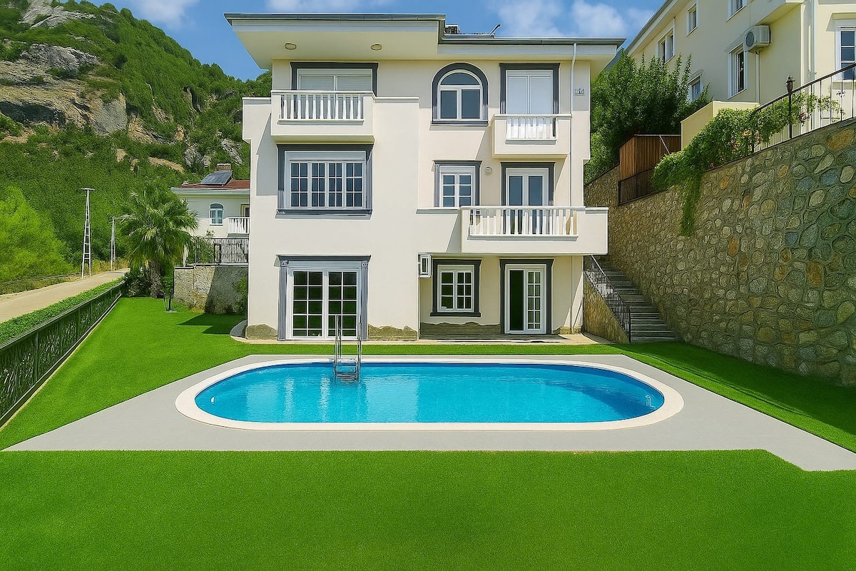 A modern villa is situated against a lush green hillside, featuring a private pool surrounded by well-maintained grass. Large windows offer natural light inside, and a balcony provides outdoor seating with views of the surrounding landscape.