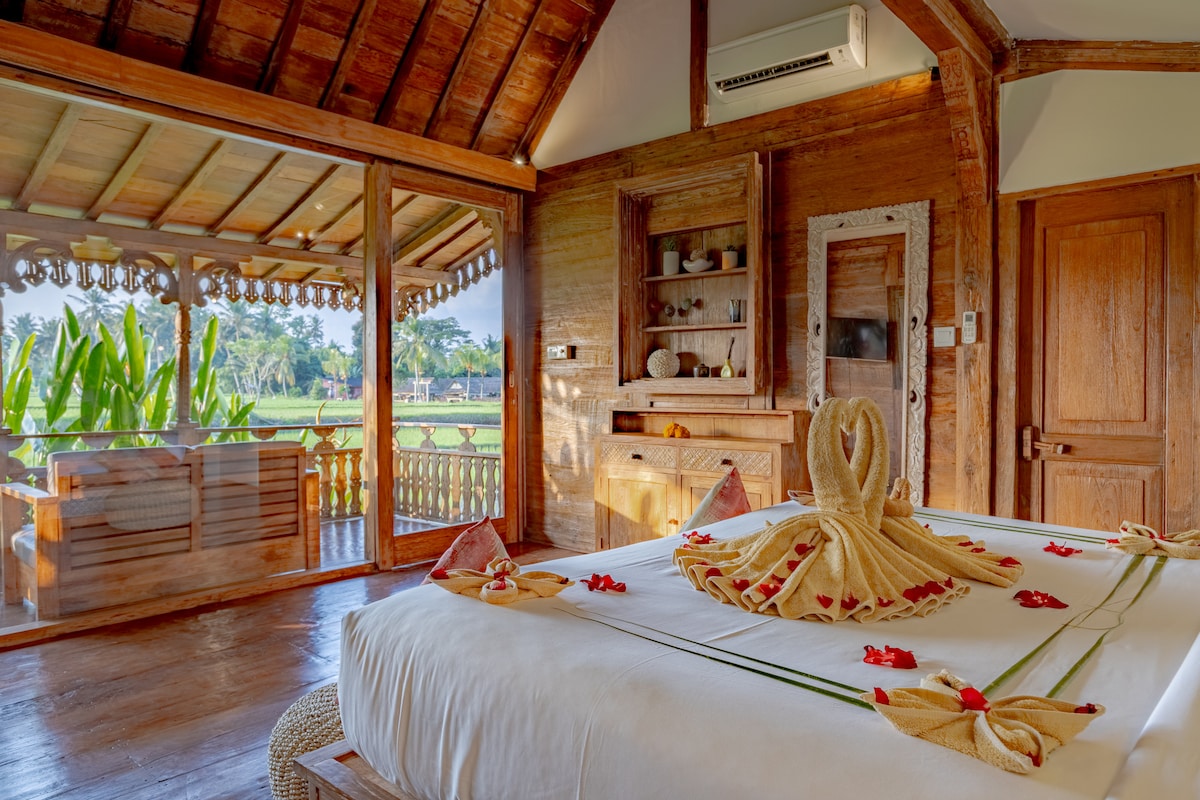 The bedroom features a spacious bed adorned with decorative towels and flower petals. Natural light filters in through the open sliding door, leading to a private patio overlooking lush greenery. Wooden accents and furnishings add warmth to the serene interior.