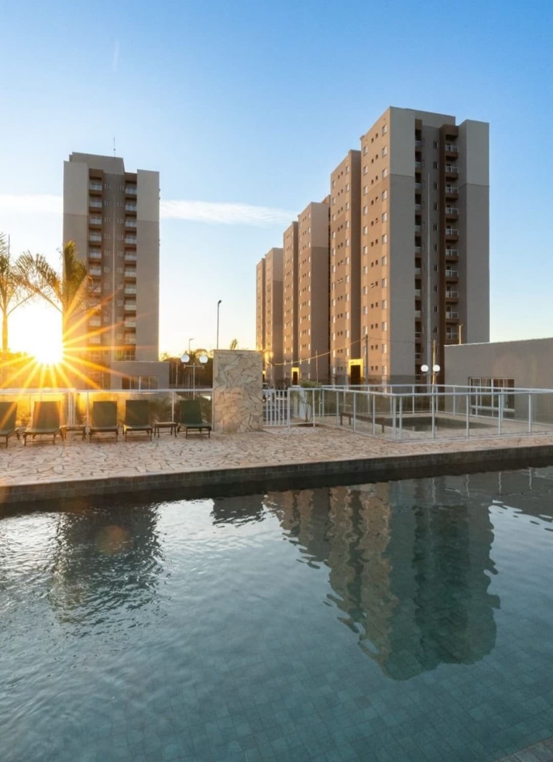 The outdoor area features a swimming pool reflecting the sunlight, surrounded by lounge chairs. Two modern residential buildings are visible in the background, along with palm trees that add to the scene's tranquility during the evening light.