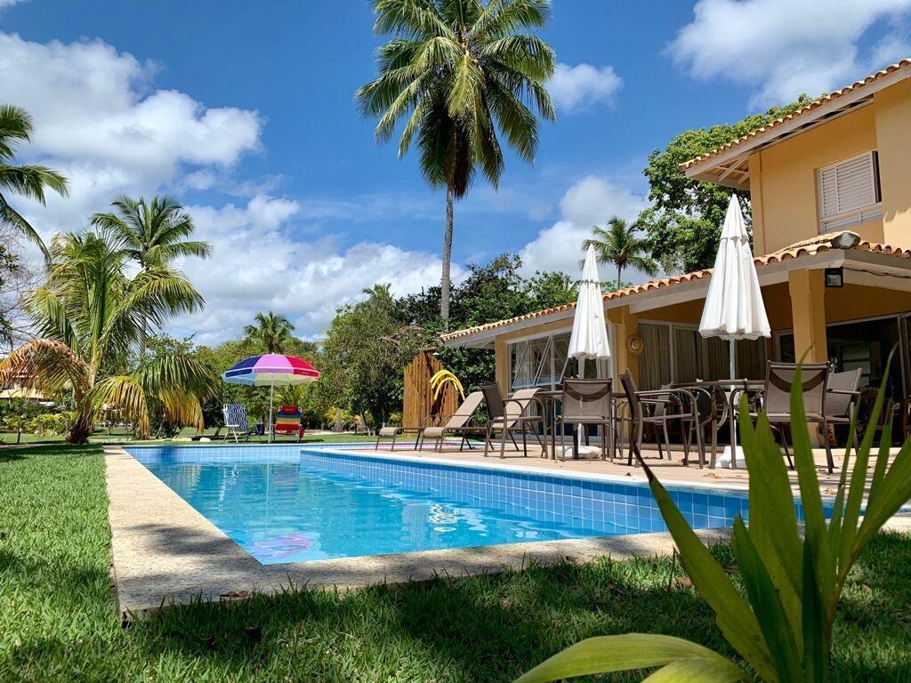 A serene outdoor view showcases a tranquil pool surrounded by lush greenery. Poolside seating is arranged under shade umbrellas, while palm trees and bright blue skies create a refreshing atmosphere. The façade of the house is visible, blending harmoniously into the landscape.