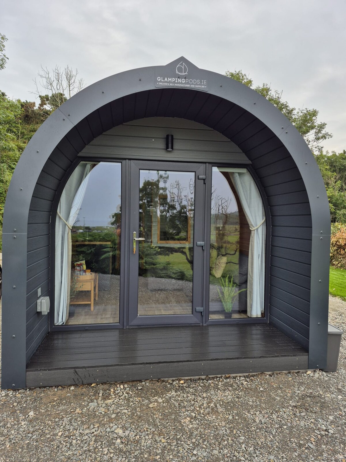 A uniquely shaped glamping pod features a rounded façade with large glass doors framed by elegant curtains. The entrance opens onto a wooden deck, surrounded by greenery. A small potted plant enhances the entrance, adding a touch of nature to the setting.