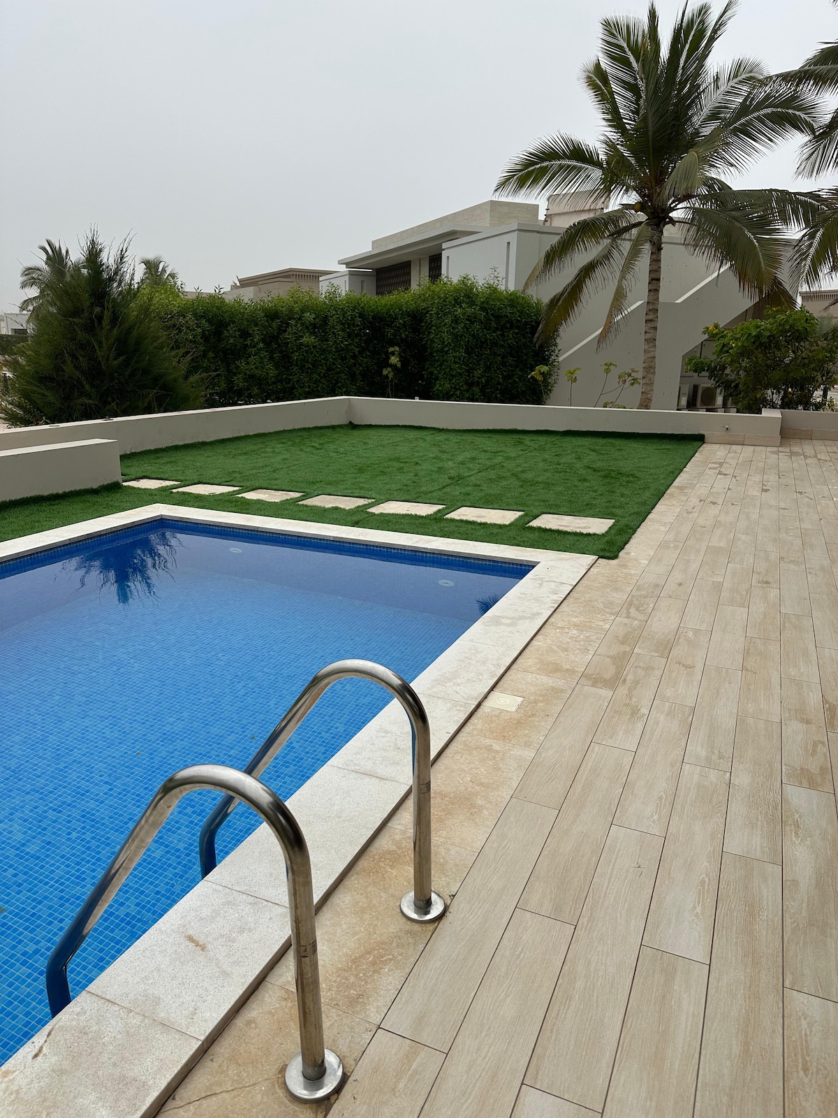 A rectangular pool, with steps leading into the water, is surrounded by a deck made of light-colored tiles. Lush green grass and stone pathways are visible along one side, with a tall palm tree providing natural shade in the background.