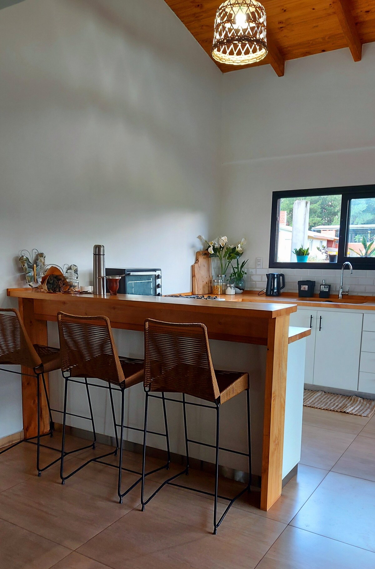 A modern kitchen area features a sleek wooden bar with three tall stools. Natural light pours in through a nearby window, illuminating the countertop adorned with small plants and kitchen appliances. Wooden beams and a wicker pendant light add a touch of warmth to the space.
