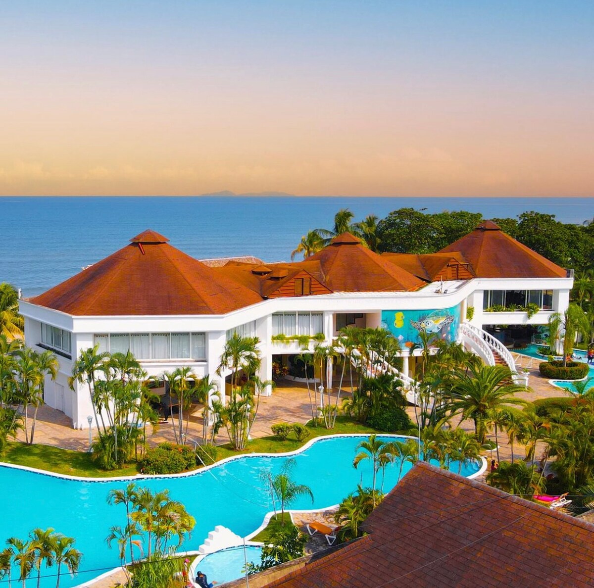 The villa is situated overlooking a sprawling resort pool and the ocean, framed by lush greenery and palm trees. The architectural design features a modern style with a distinct roof shape and expansive windows, allowing for abundant natural light.