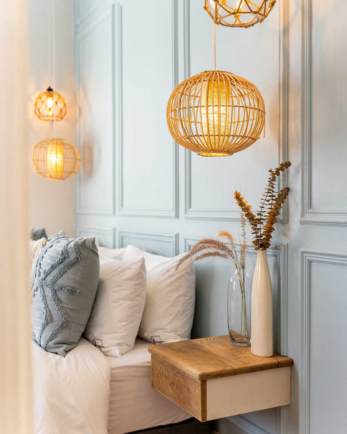 A cozy corner of the home features a light blue wall with decorative molding, illuminated by woven pendant lights. A bedside table made of natural wood holds a slender vase containing dried foliage, while plush white pillows are arranged on a spacious bed.