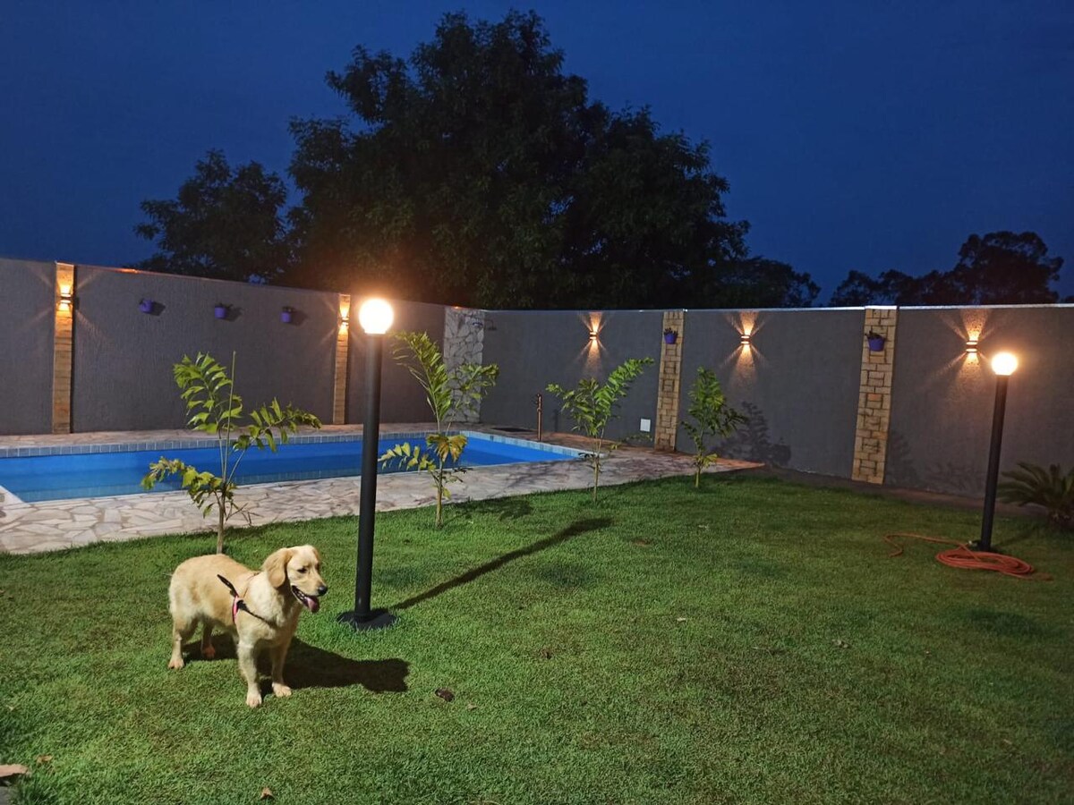 A well-maintained backyard is highlighted by a tranquil swimming pool surrounded by neatly cut grass. Decorative lights illuminate the area, creating a warm ambiance. A golden retriever stands on the grass, adding a lively presence to the scene. A fenced perimeter is visible in the background.