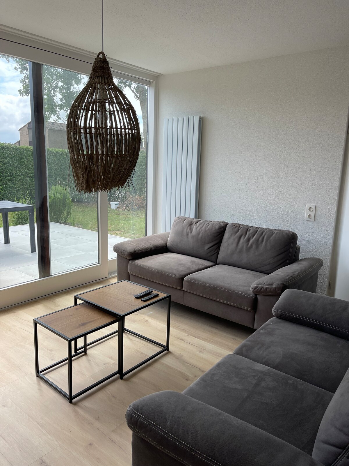 A comfortable living area features a gray sofa and an armchair, arranged around a small wooden coffee table. Sunlight enters through large glass doors, illuminating the space and revealing views of the outdoor area. A woven pendant light adds texture to the ceiling.