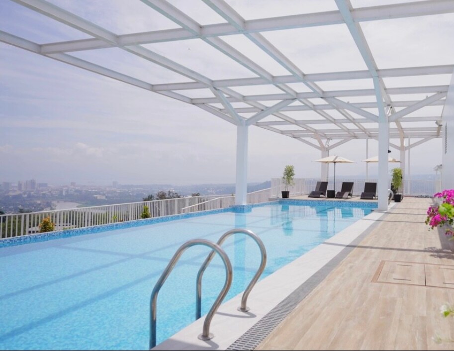 A rooftop pool area is featured, showcasing an inviting blue pool under a white pergola. Comfortable lounge chairs are positioned nearby, offering a panoramic view of the city skyline. Flowering plants add color to the surrounding space, enhancing the serene environment.