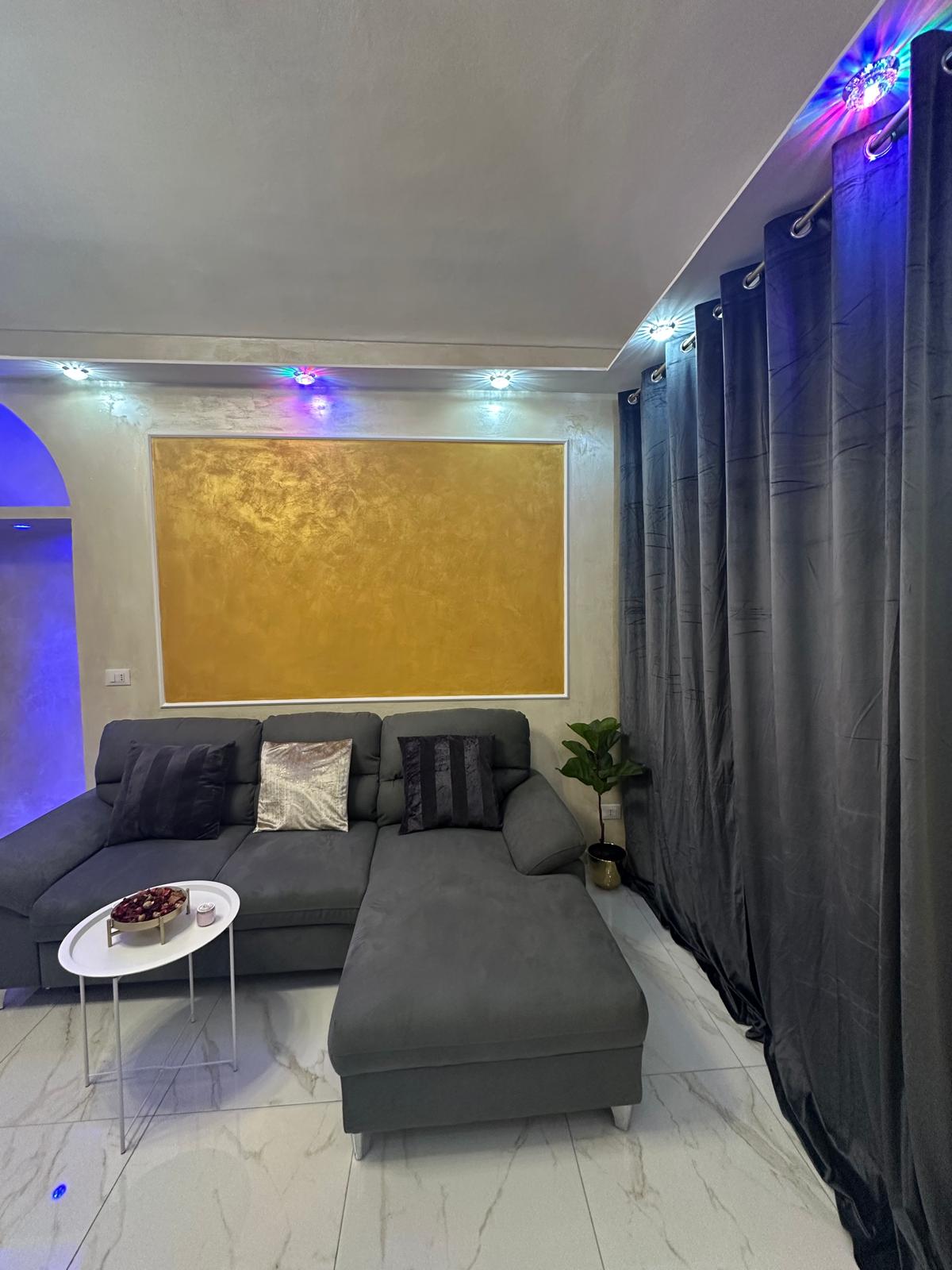 A sectional sofa is situated in a corner, complemented by decorative cushions in varying textures. A small round table is positioned nearby, adorned with a bowl of snacks. The wall features a gold accent above the sofa, illuminated by soft, multicolored lighting.