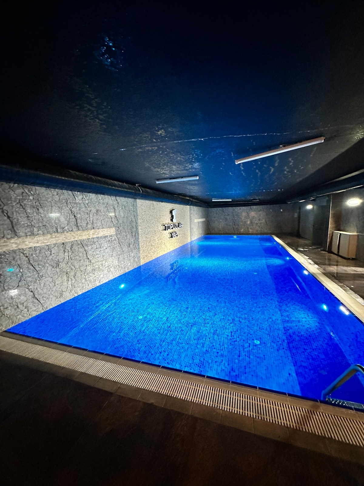 An indoor swimming pool is presented, featuring a vibrant blue hue. The sleek, marble walls offer a modern touch. Soft lighting enhances the serene atmosphere, while the pool area is spacious, allowing for comfortable movement around the water's edge.
