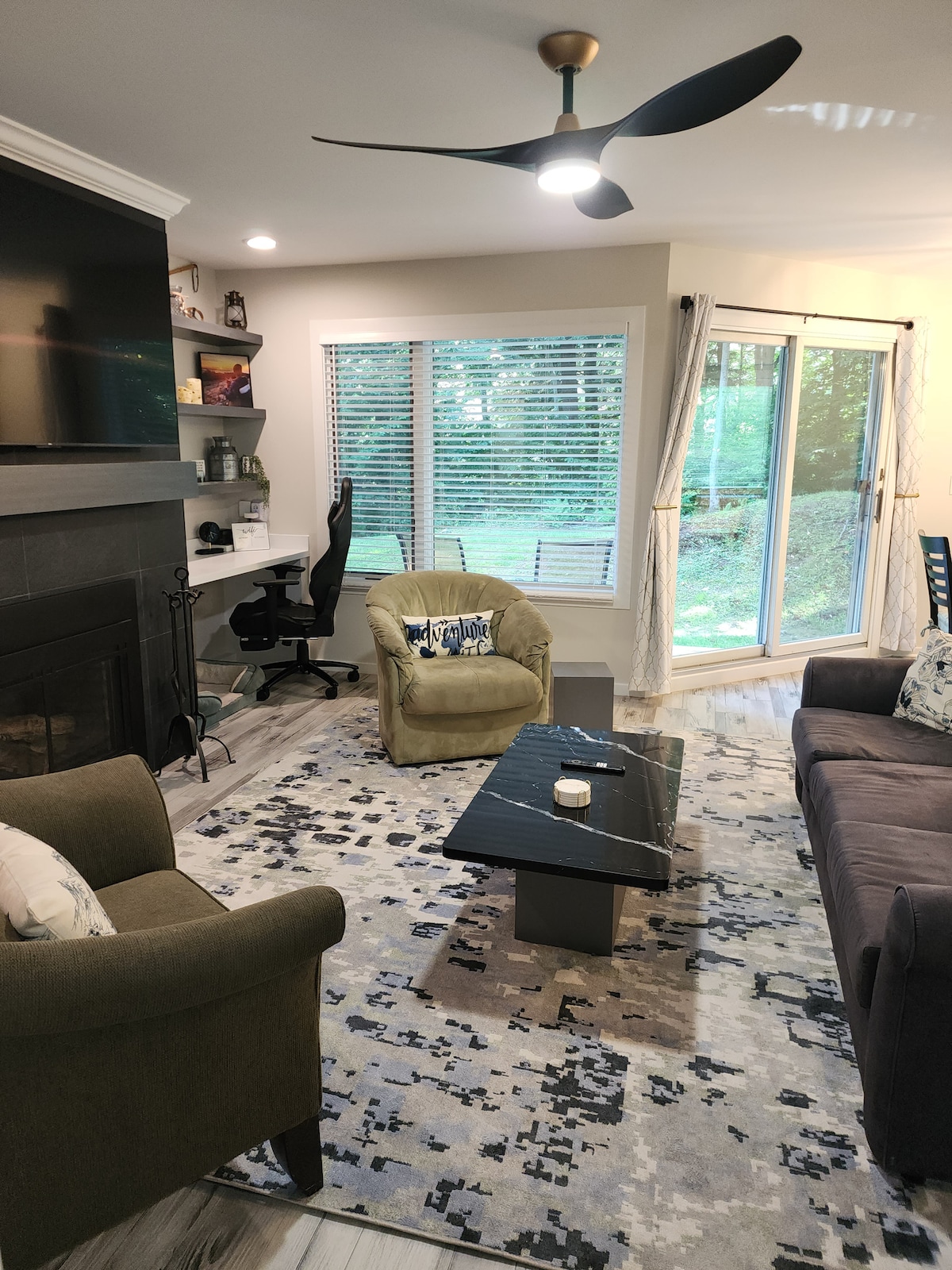 The cozy living area is equipped with a gas fireplace and features a spacious dark sofa and two armchairs. A small dining table is centered on a patterned rug, with soft light coming through large windows overlooking the outdoor greenery. A designated workspace is visible.