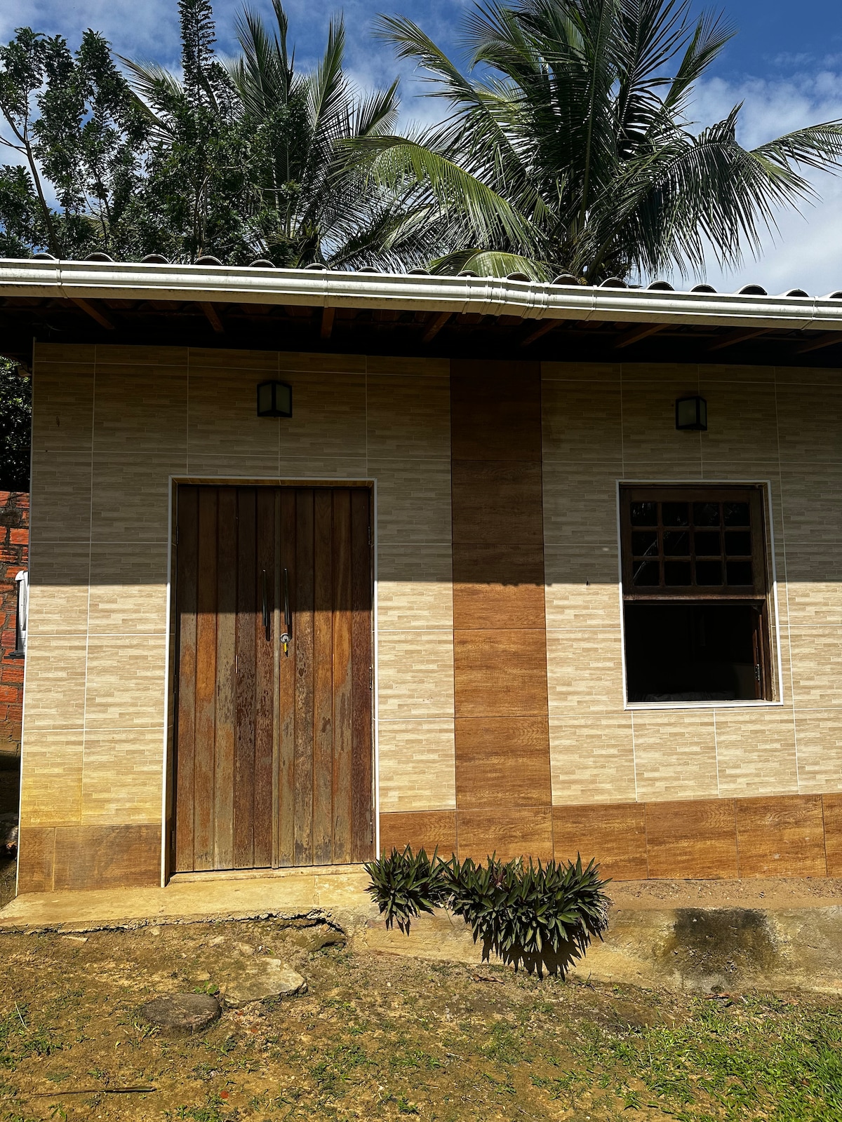 An exterior view of a modest house is showcased, featuring a wooden door and a single window. The facade is adorned with patterned tiles in light and earthy tones. Lush green plants are visible in front, with palm trees seen in the background under a clear blue sky.