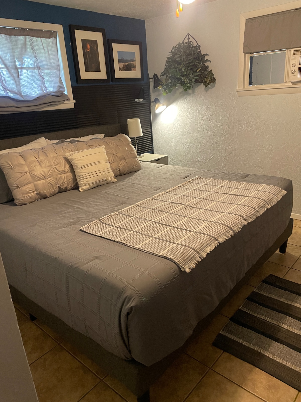 A cozy sleeping space features a spacious bed dressed in soft gray linens. A checkered throw blanket is neatly placed at the foot of the bed. Wall art adds character, while natural light enters through a window, complemented by a soft window treatment.