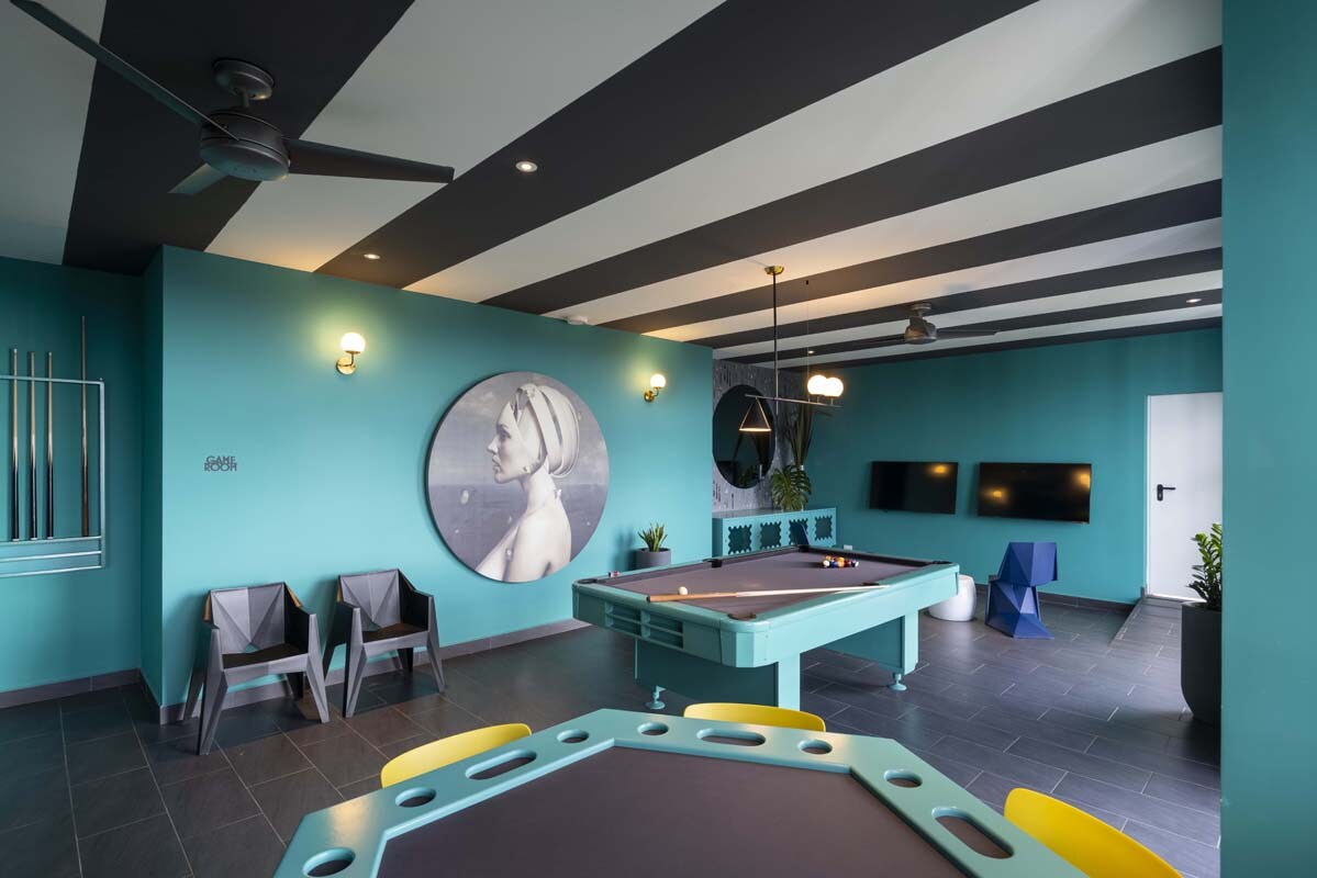 A game room is presented with a pool table as the focal point, surrounded by vibrant teal walls and striped ceilings. Seating arrangements include colorful chairs and outdoor-style lounge chairs. Two flat-screen TVs are mounted on the wall, providing entertainment options within the spacious area.