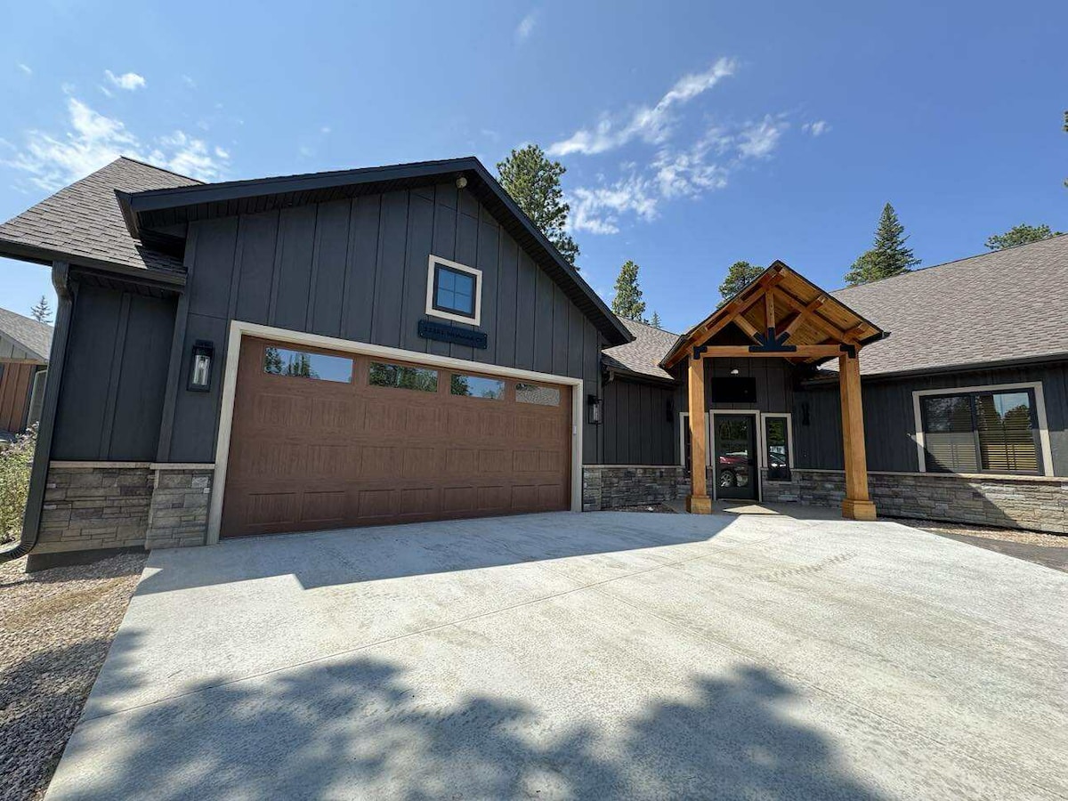 A modern home exterior is displayed, featuring dark wood siding and a large stone entry. A spacious driveway is visible, leading to a two-car garage with a brown door. The covered entryway showcases wooden beams, surrounded by trees and a clear blue sky.