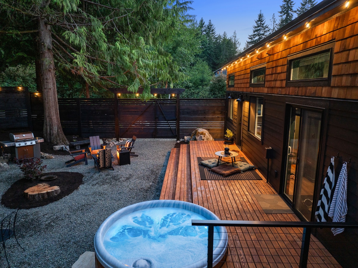 An inviting outdoor space features a hot tub surrounded by a wooden deck, with a fire pit and two lounge chairs arranged nearby. Soft lighting accentuates the area, while tall cedar trees provide natural privacy. A barbecue grill is positioned to the side, enhancing outdoor dining options.