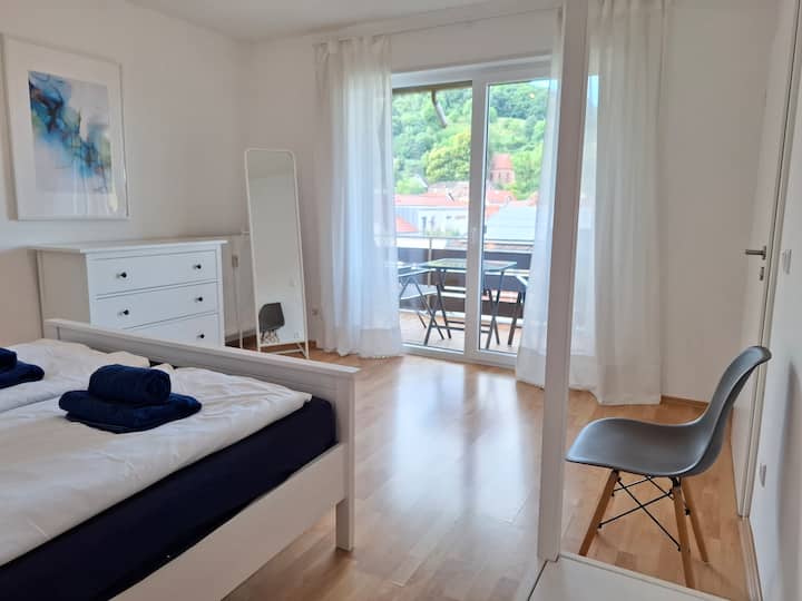 明亮宽敞的双人卧室，配有梳妆台、桌子、镜子和遮光帘。 可直接进入阳台，可欣赏莱恩斯韦勒屋顶的壮丽景色。