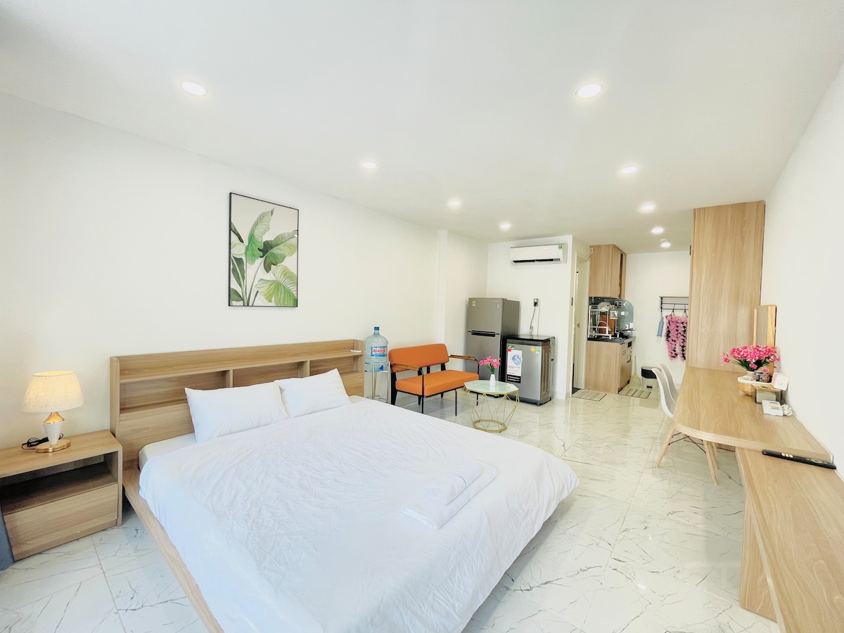 A spacious studio apartment features a large bed with clean linens positioned against a wooden headboard. A small lamp on a bedside table casts a soft glow. An orange seating area adds a pop of color, while a work desk stands against the wall, equipped with a chair. A kitchenette is visible in the background.