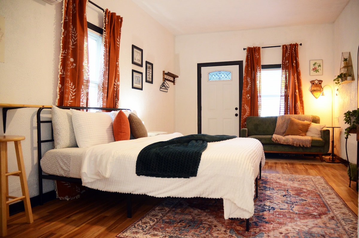 The studio features a comfortable king-size bed with soft linens, paired with accent pillows. A cozy seating area is visible with green upholstery, complemented by warm curtains and a colorful area rug. Natural light enters through windows, adding to the inviting ambiance.