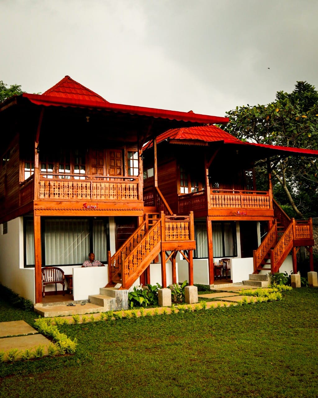 A charming two-story wooden villa is depicted, featuring a red roof and spacious balconies. The ground is adorned with lush green grass and neatly arranged plants, while stone pathways lead to the entrance. A person can be seen sitting on a chair, enjoying the serene surroundings.