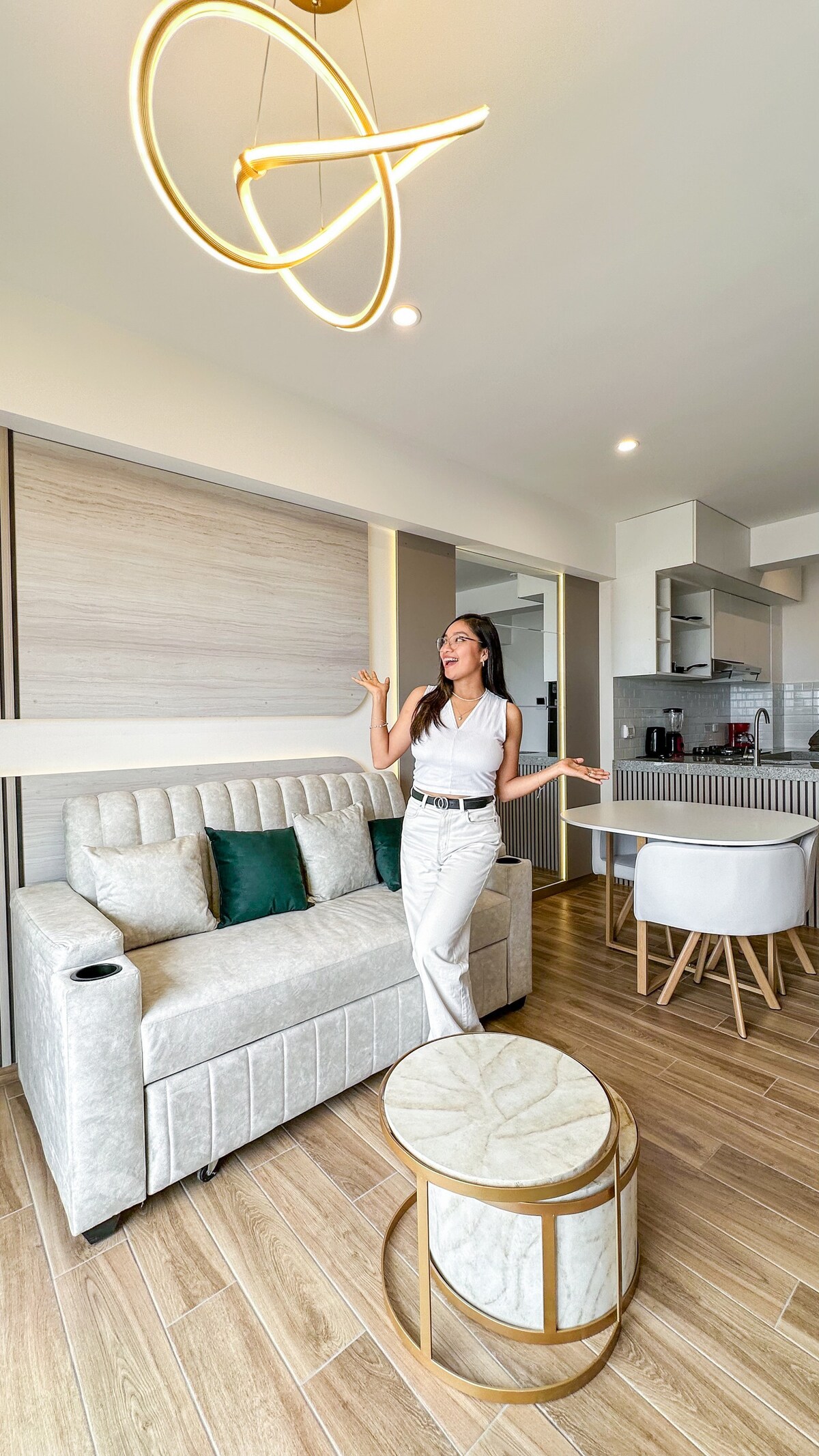 A modern living area features a comfortable sofa adorned with green cushions. A stylish coffee table with a marble top is positioned in front. The space is illuminated by a contemporary light fixture overhead, and a glimpse of the kitchen area is visible in the background.