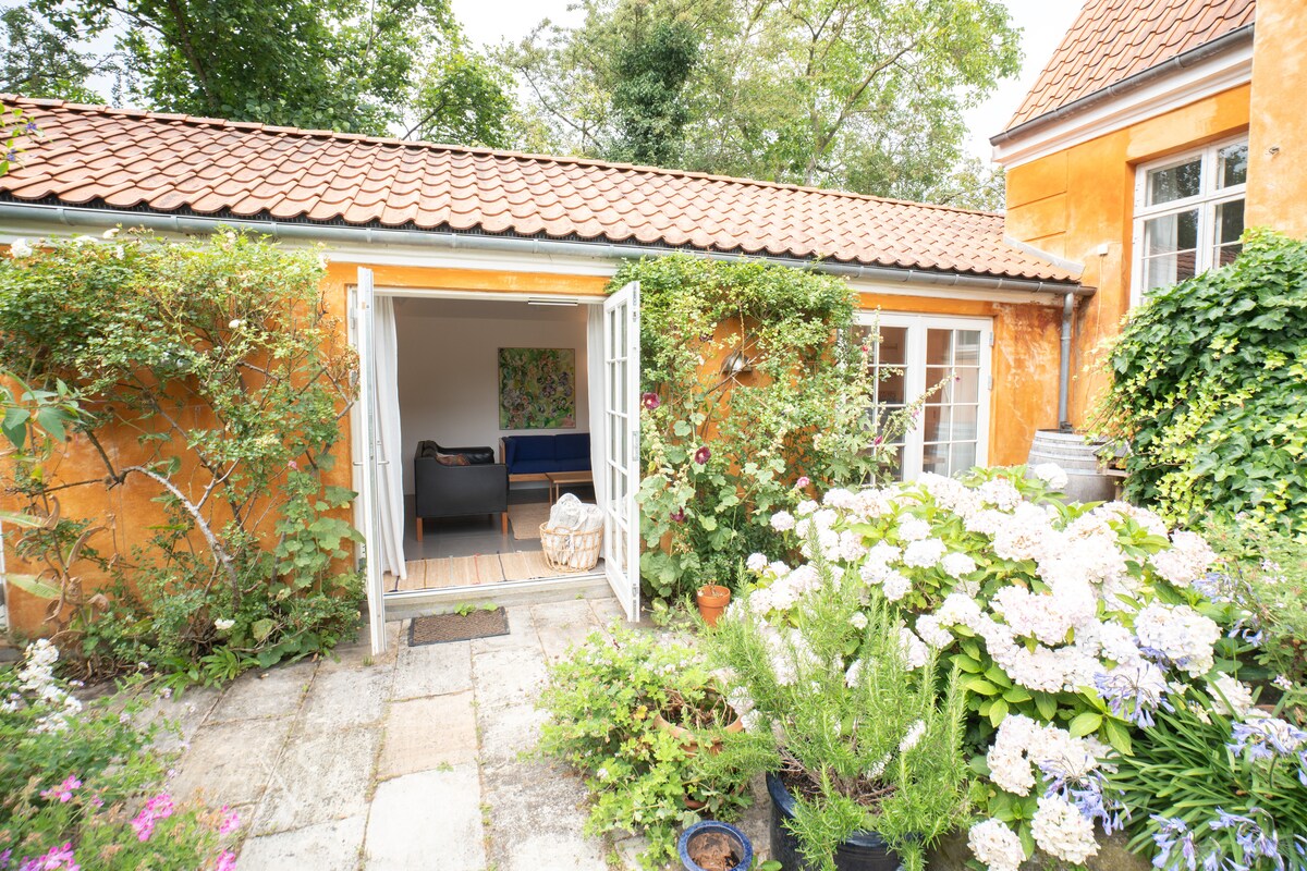 A charming entrance to a guesthouse is framed by vibrant greenery and flowering plants. Double doors are opened to reveal a bright interior, while a cozy seating area with a blue couch is visible. The warm, ochre exterior contrasts with the lush garden.