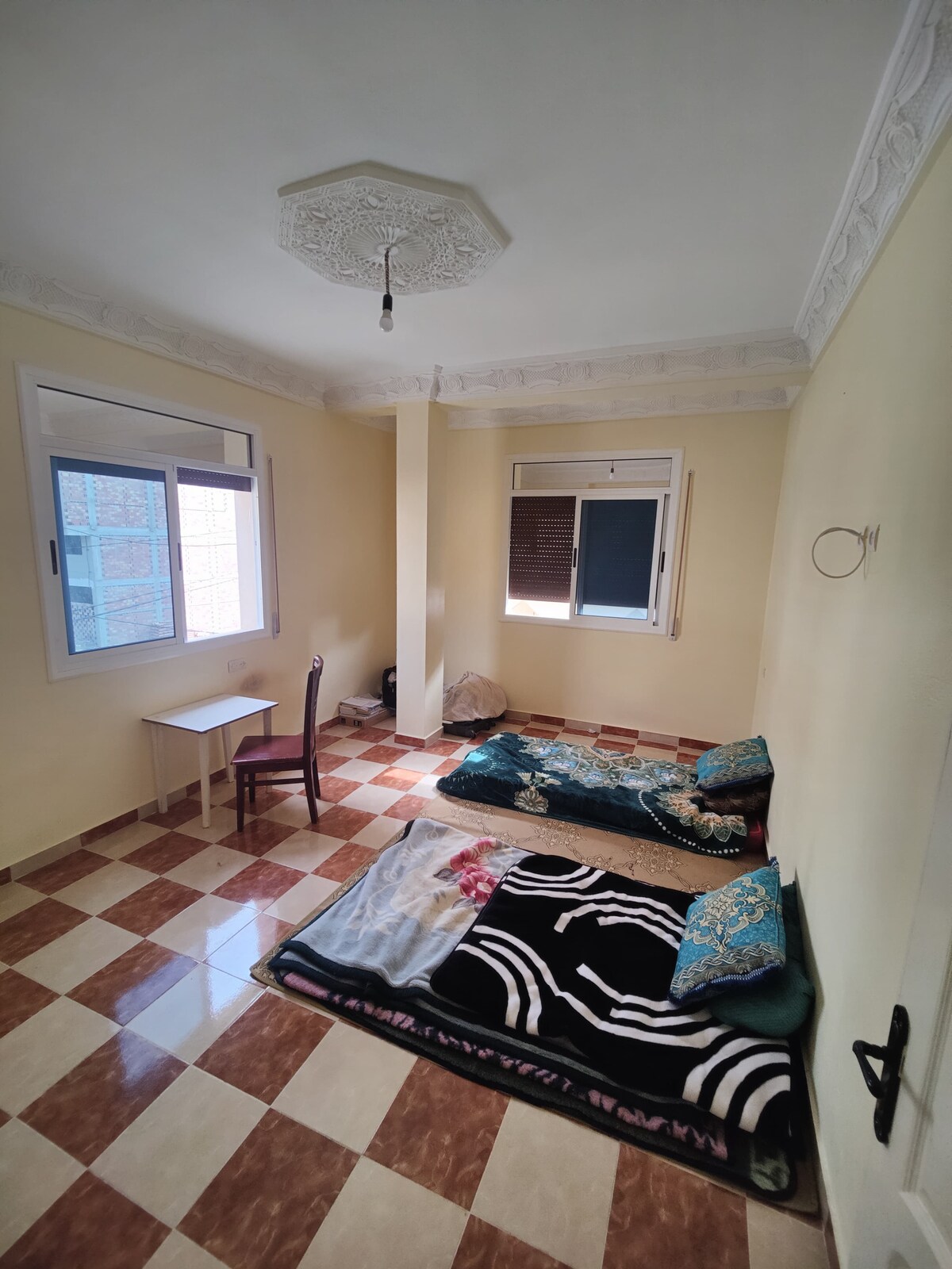 A bright room features patterned tile flooring, with two spacious sleeping areas arranged on the ground. A small table is positioned against the wall next to a chair. Light enters through a window, illuminating the neutral-colored walls and ceiling design.