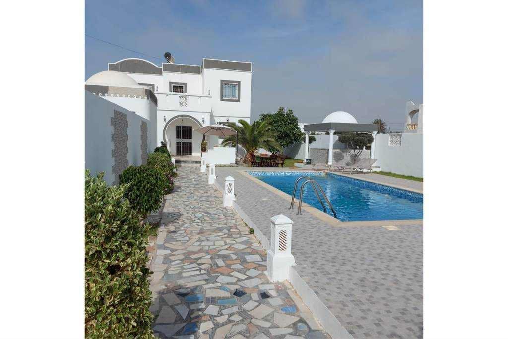 A charming villa is surrounded by a stone pathway and lush greenery, leading to a private swimming pool. The pool area features an umbrella-shaded seating space and a landscaped garden. A white wall encloses the property, contributing to a sense of privacy.
