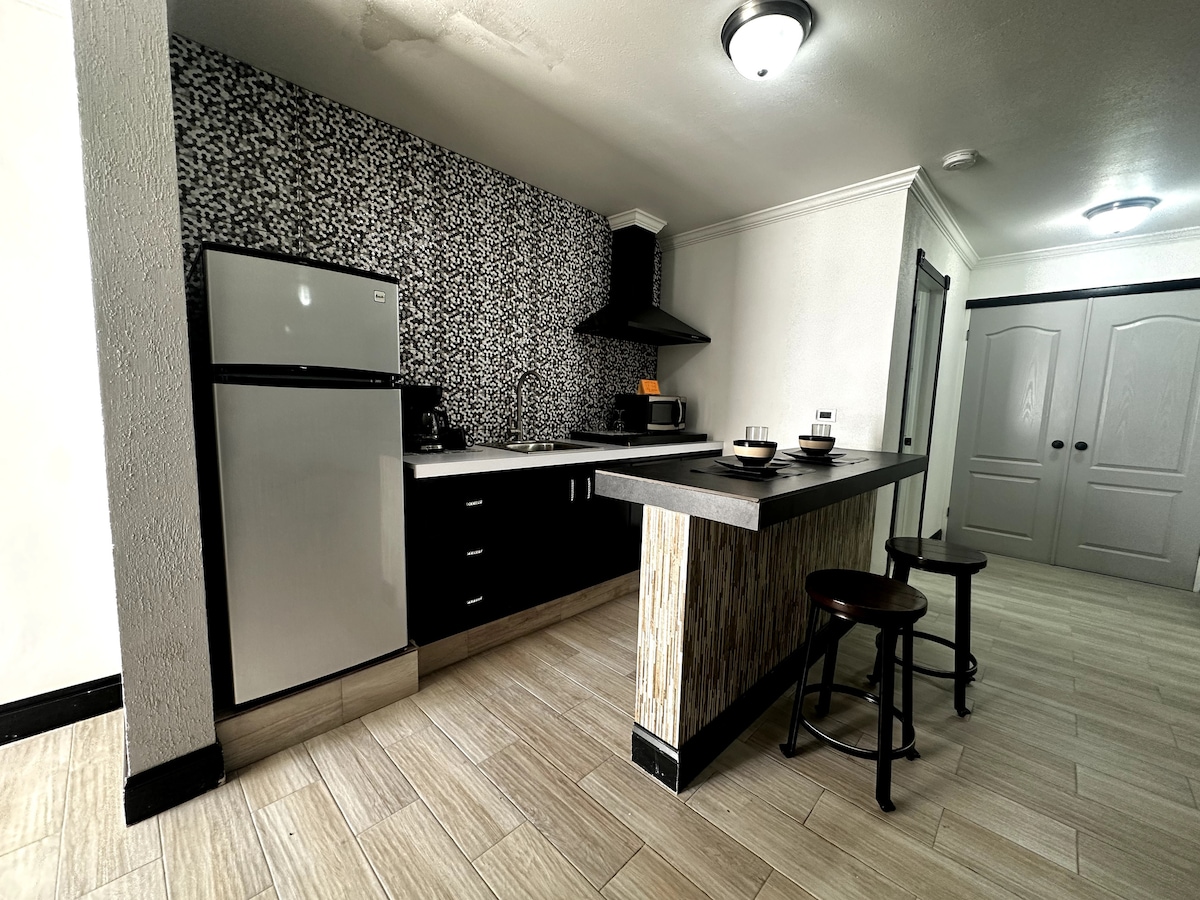 A modern kitchen is featured with sleek black cabinetry and a contrasting patterned wall. Stainless steel appliances are visible, including a refrigerator and stove. A breakfast bar with two black stools invites casual dining. Soft, neutral lighting enhances the clean lines and contemporary design.