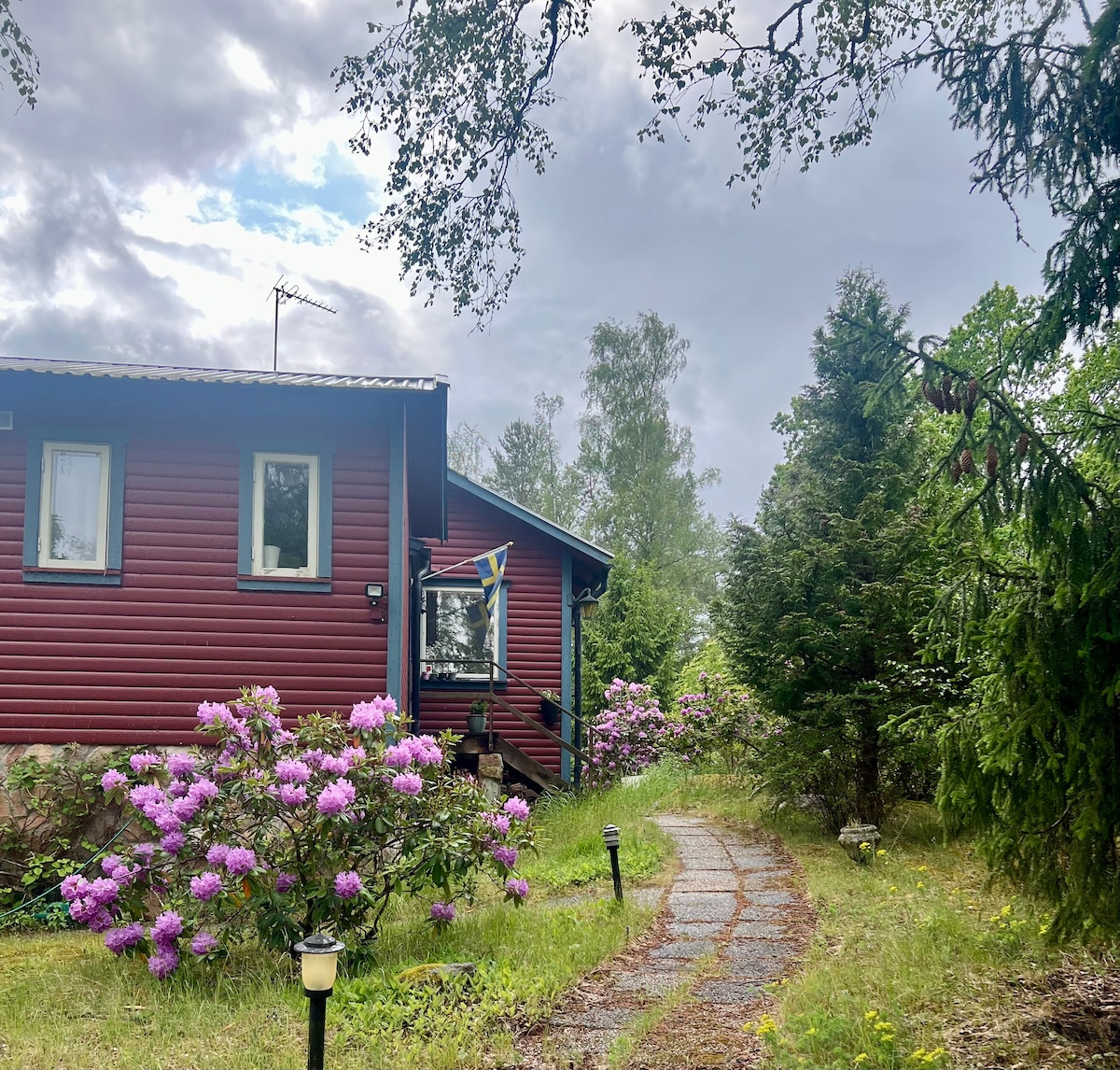 A charming red cottage with a front porch is set amidst greenery. A pathway lined with stones leads to the entrance, flanked by blooming pink rhododendrons. Lush trees provide a natural border, while soft lighting fixtures illuminate the walkway.