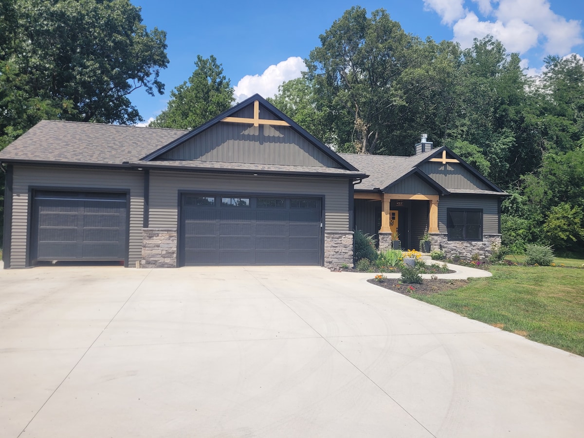 The exterior of the property is showcased, featuring a modern design with a combination of dark siding and stone accents. A welcoming entrance is visible, flanked by landscaped flower beds. Two garage doors are prominent, providing ample parking space on a paved driveway.