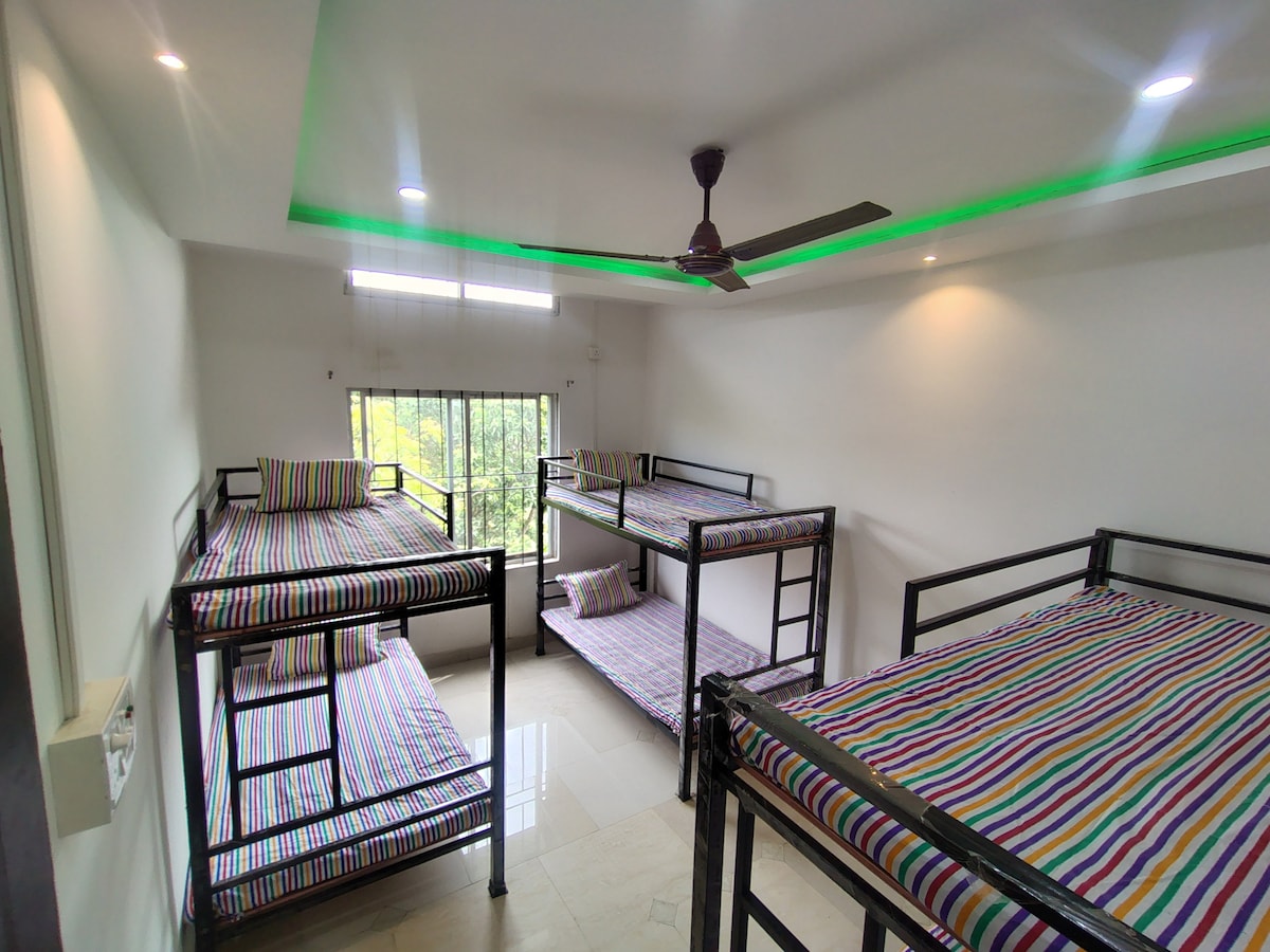 A dormitory room features multiple metal bunk beds, each covered with striped bedding. Natural light filters through the large window, enhancing the clean and airy feel of the space. A ceiling fan is positioned above, contributing to a comfortable atmosphere.