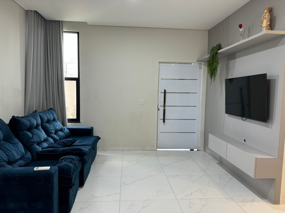 A spacious living area features a plush blue sofa arranged around a flat-screen television mounted on a clean white wall. Natural light filters through a large window beside the door, which is framed by neutral-toned curtains. The glossy floor tiles enhance the room's brightness.