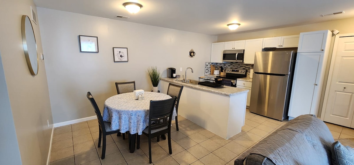 A kitchen and dining area display a light-colored tile floor. A round dining table with six chairs is set with a tablecloth. Appliances include a stainless-steel refrigerator and oven, while a sink and countertop offer ample space. Subtle wall décor adds to the atmosphere.