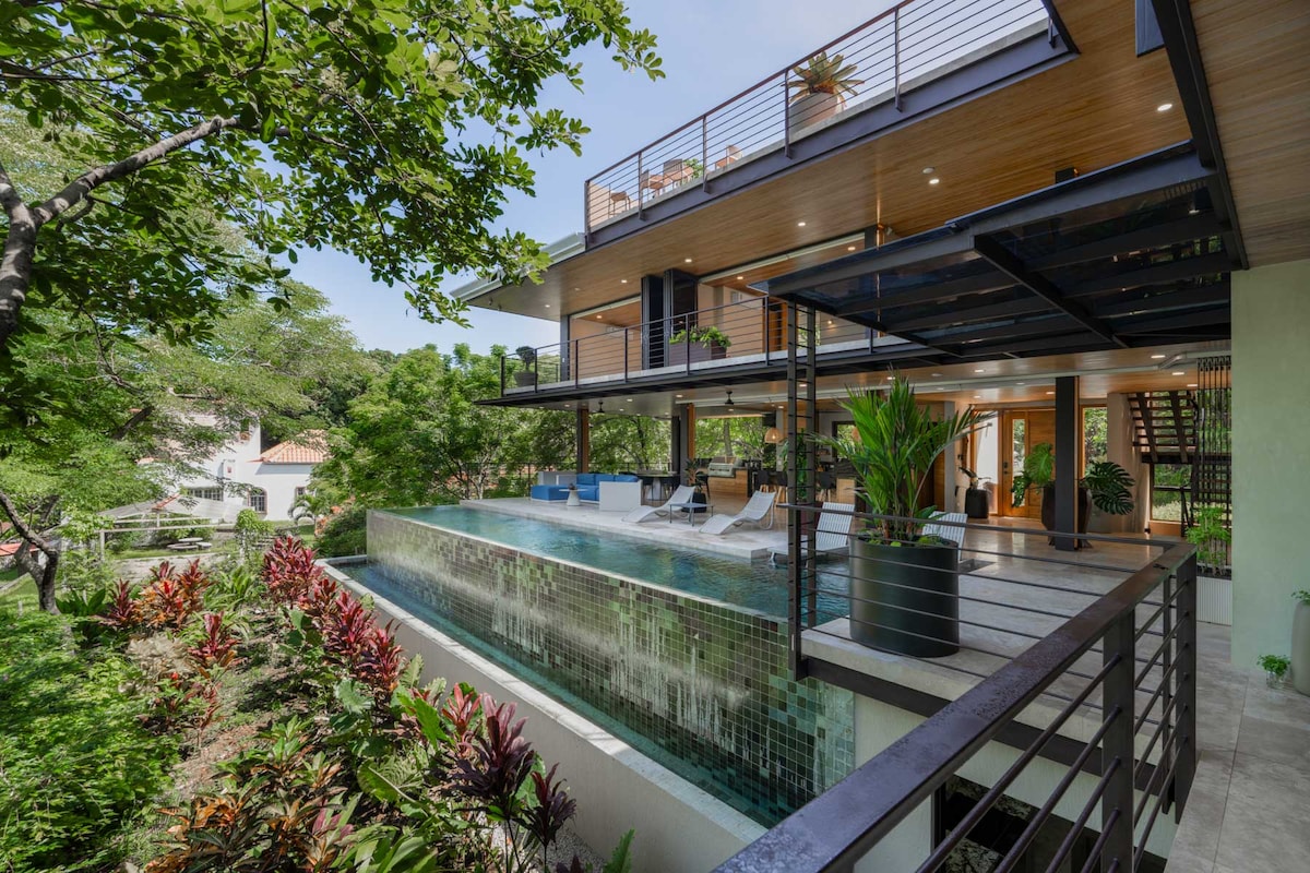 A modern home is surrounded by lush greenery, featuring a spacious terrace with lounge chairs. An infinity pool extends outward, blending into the landscape. The structure's open design allows natural light to illuminate the interior spaces, creating a seamless connection to nature.
